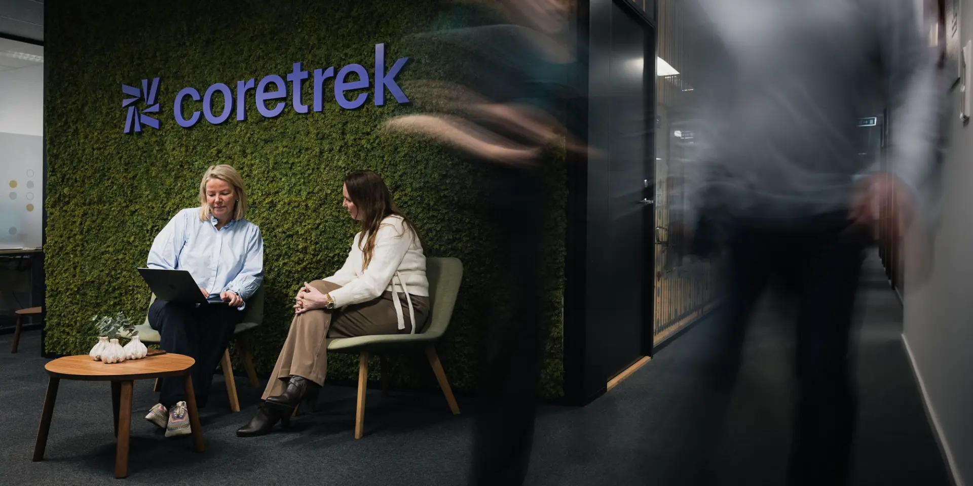 To personer sitter foran en mosekledd vegg med coretrek logo i lilla